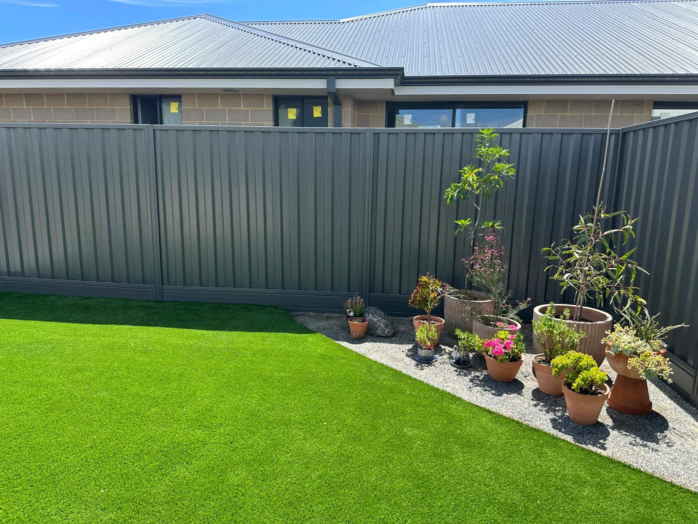 Grey Colorbond fence behind a gravel garden bed of potted plants and synthetic lawn