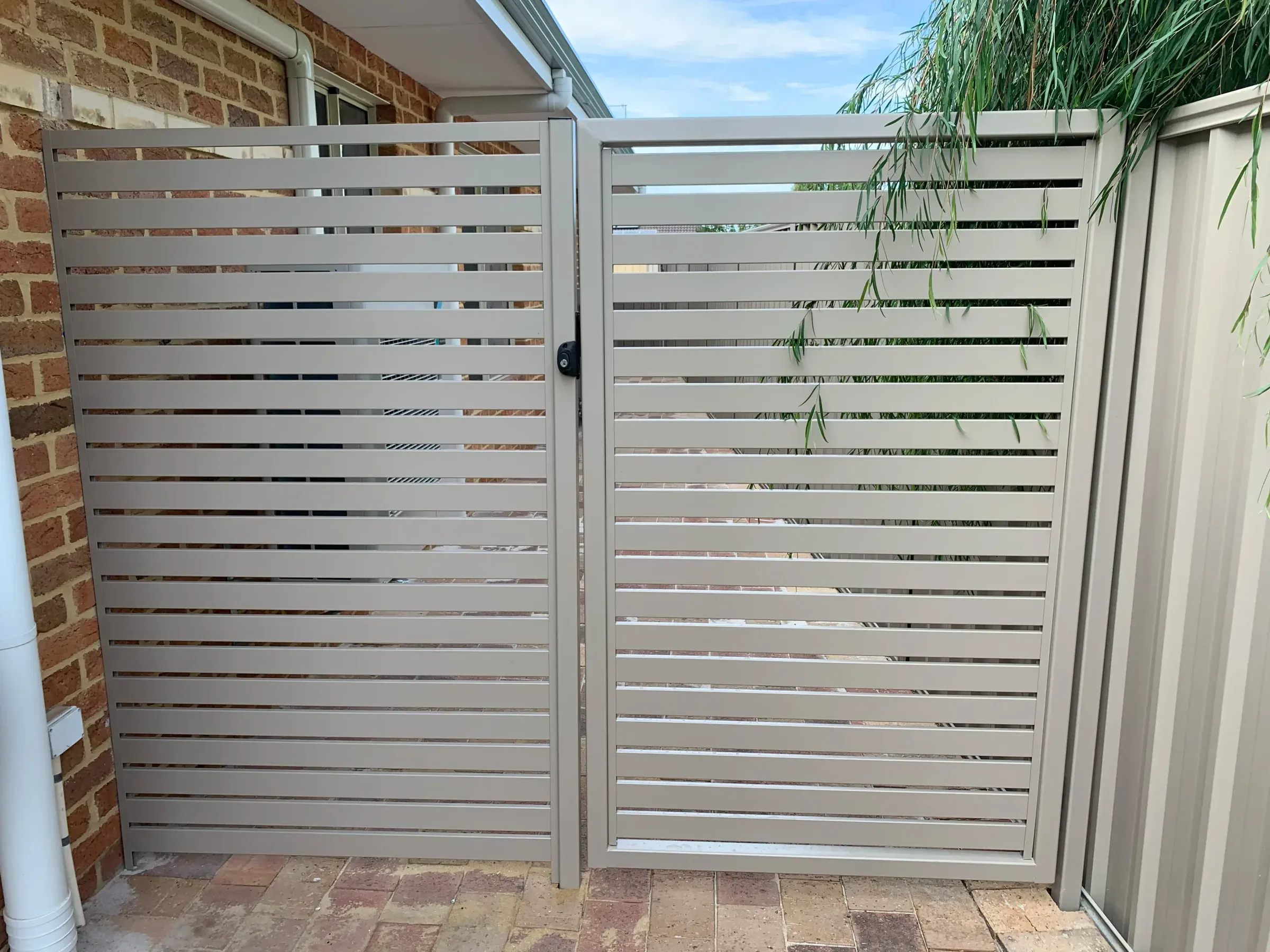 Aluminium slat double gate hung between brick piers beside a house wall