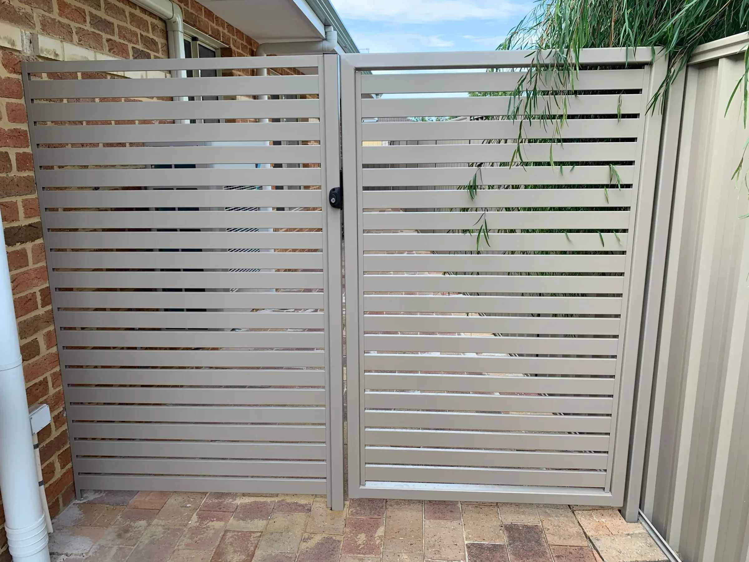 Aluminium slat pedestrian gate giving side access between a brick pier and a fence panel