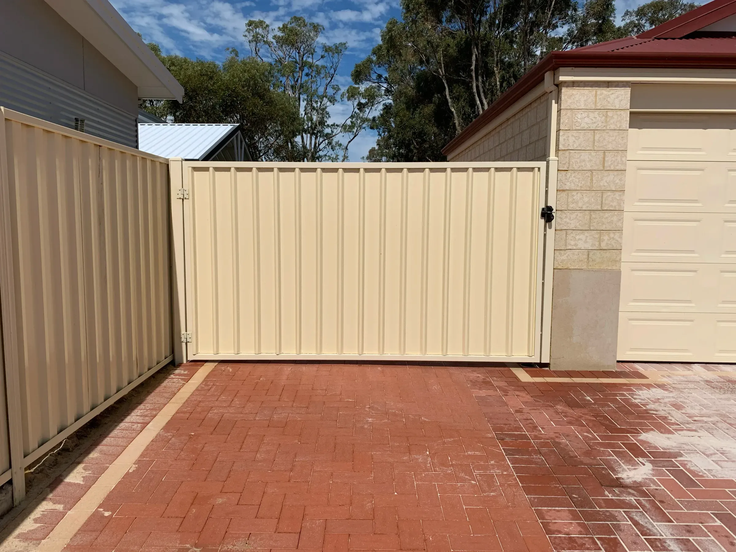 Cream Colorbond double gate closed across brick paving between two rendered walls