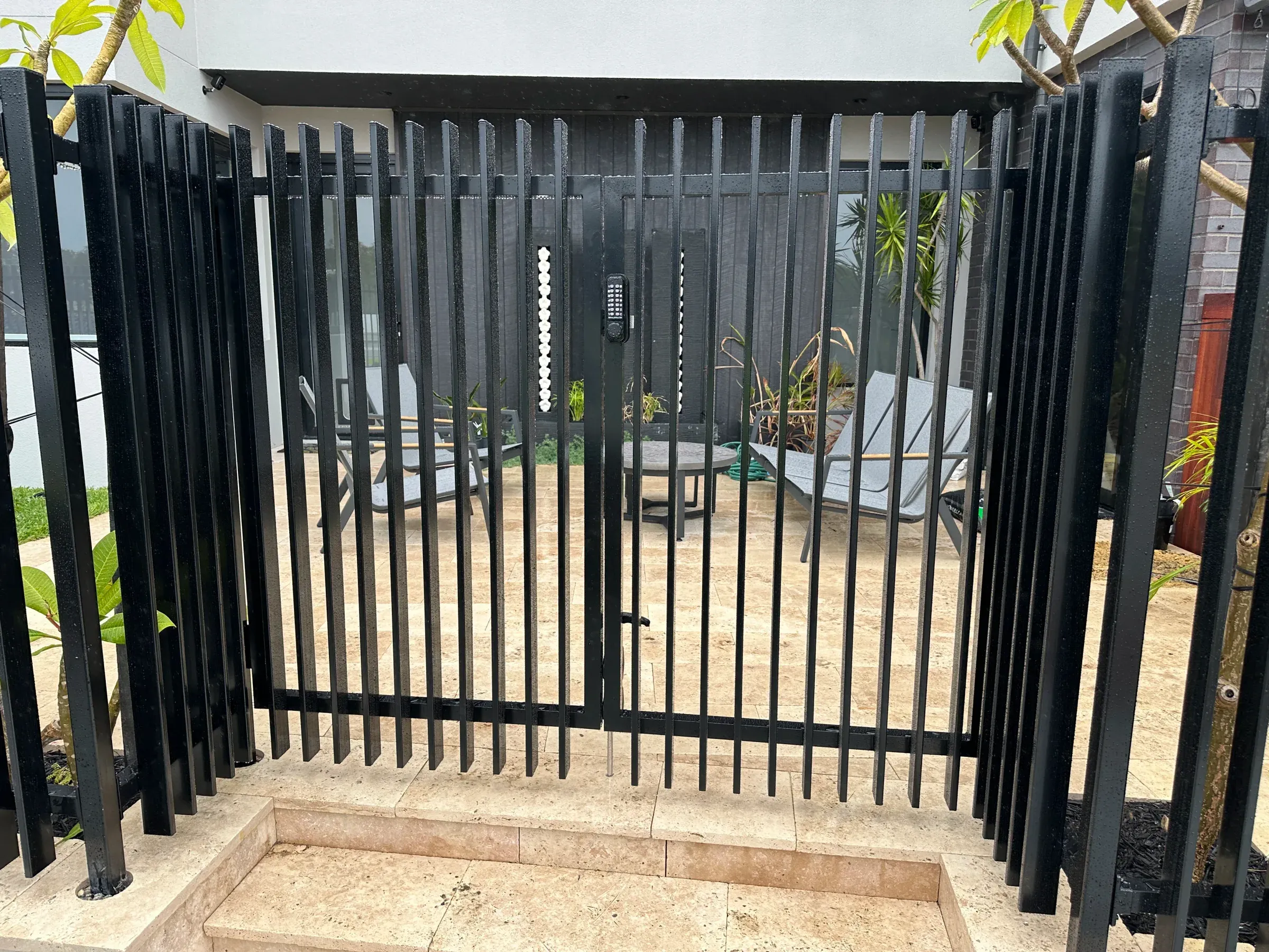 Black tubular gate and fence enclosing a paved courtyard beside a limestone wall